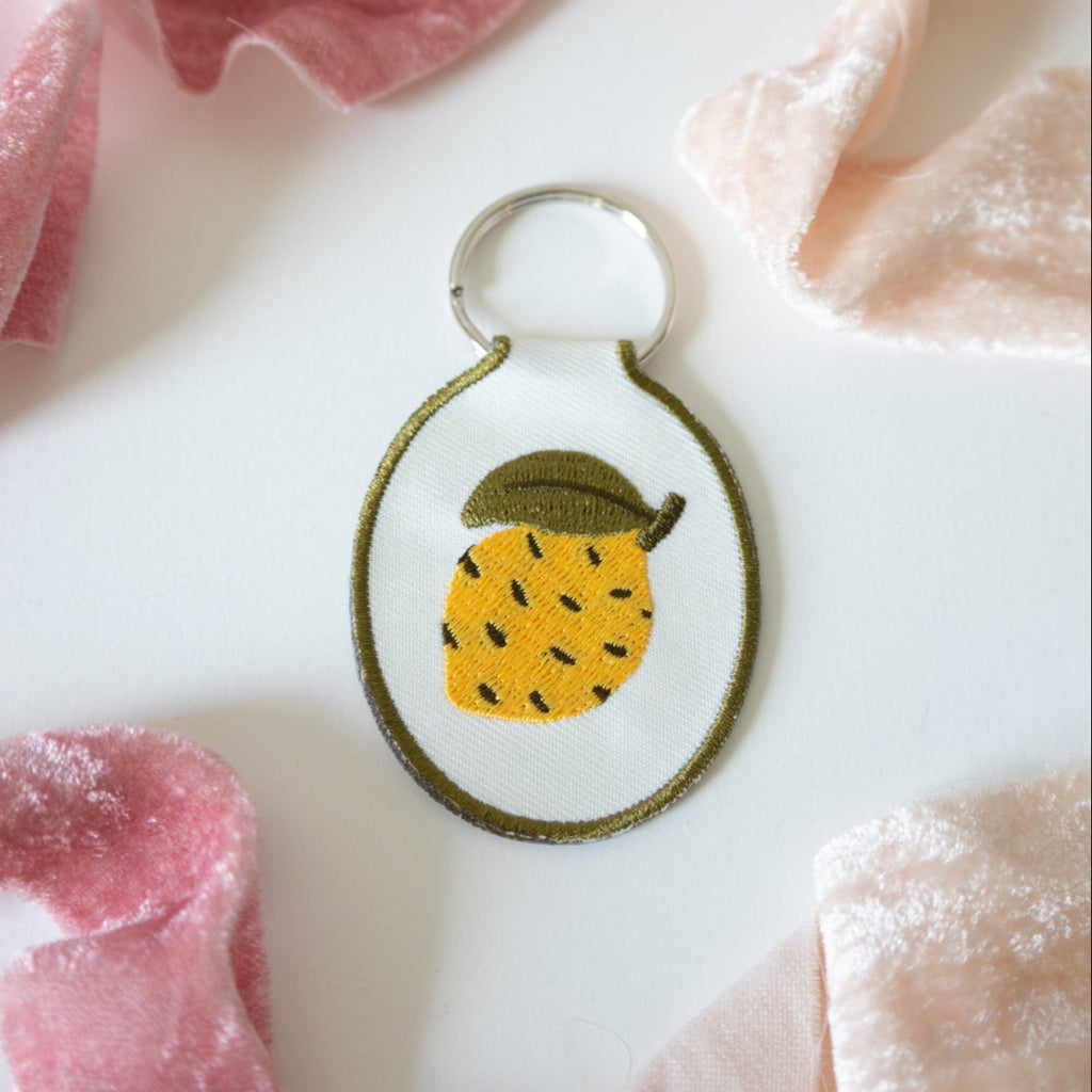 Embroidered lemon keychain sitting on a white surface with pink ribbon.