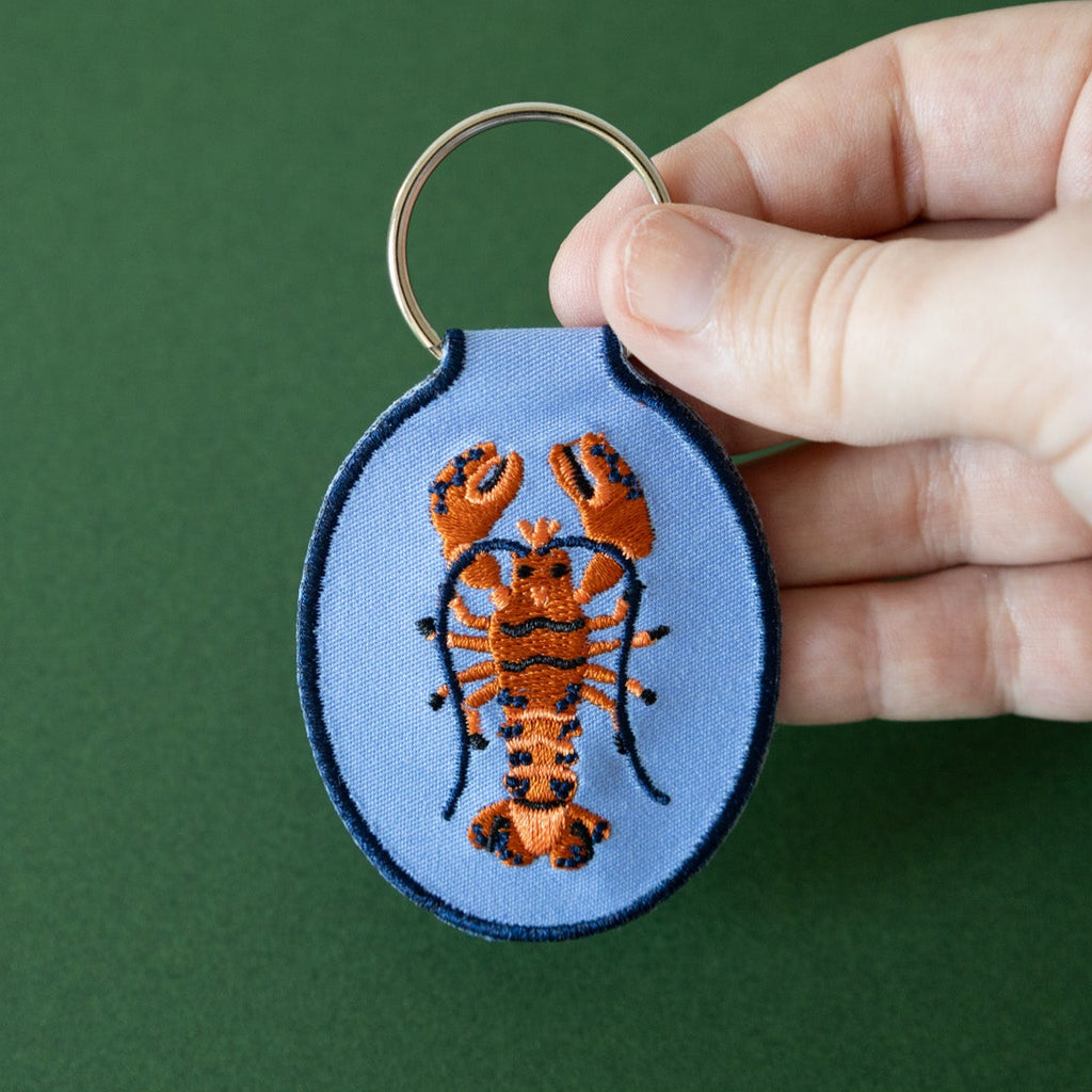 A hand holding a blue keychain with an embroidered red and blue lobster design, displayed against a green background.