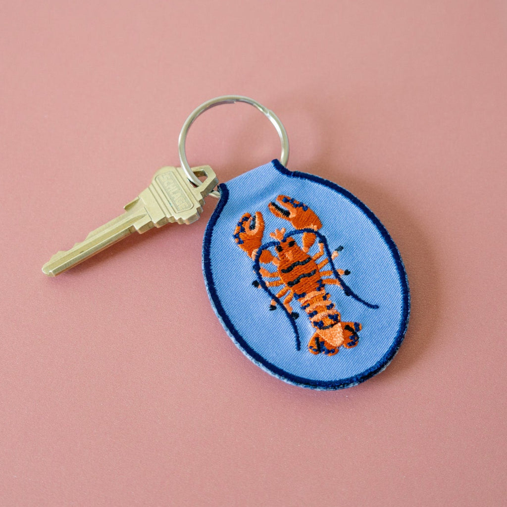 A blue keychain with an embroidered red and blue lobster design, displayed against a pink background.
