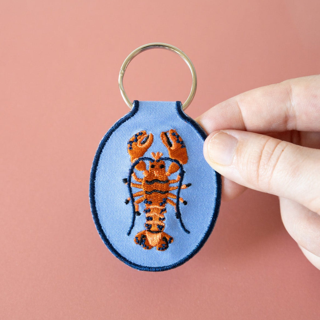 A hand holding a blue keychain with an embroidered red and blue lobster design, displayed against a pink background.