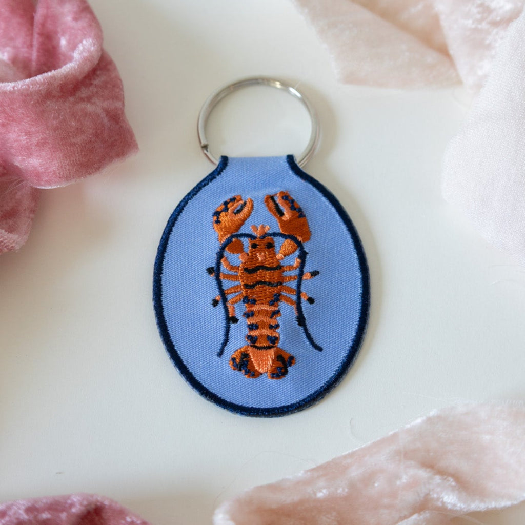 A blue keychain with an embroidered red and blue lobster design, displayed against a white background with pink ribbons.
