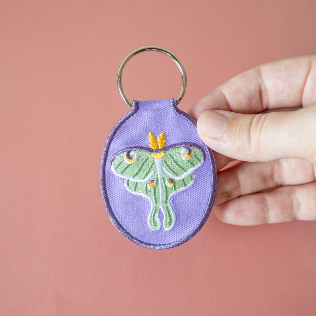 Purple keychain with embroidered luna moth design held by a hand against a pink background
