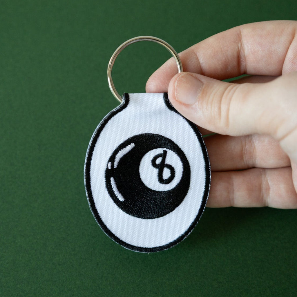 A hand holding an embroidered keychain with a white background featuring a black eight ball design, displayed on a green background.