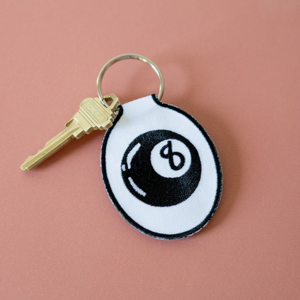 An embroidered keychain with a white background featuring a black eight ball design, displayed on a pink background.