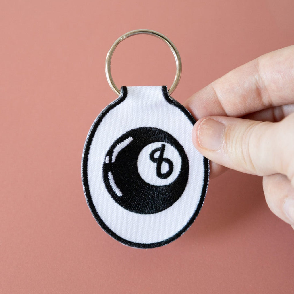 A hand holding an embroidered keychain with a white background featuring a black eight ball design, displayed on a pink background.