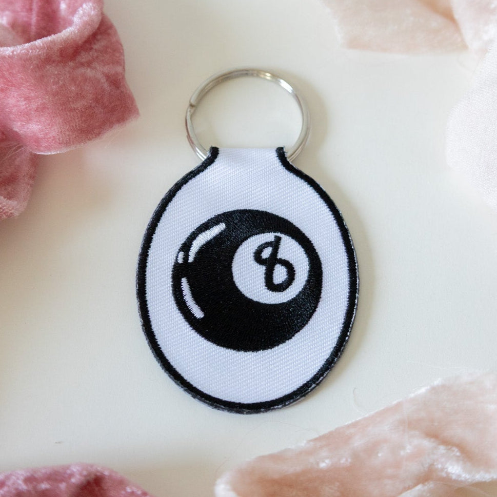 An embroidered keychain with a white background featuring a black eight ball design, displayed on a white background with pink ribbons.