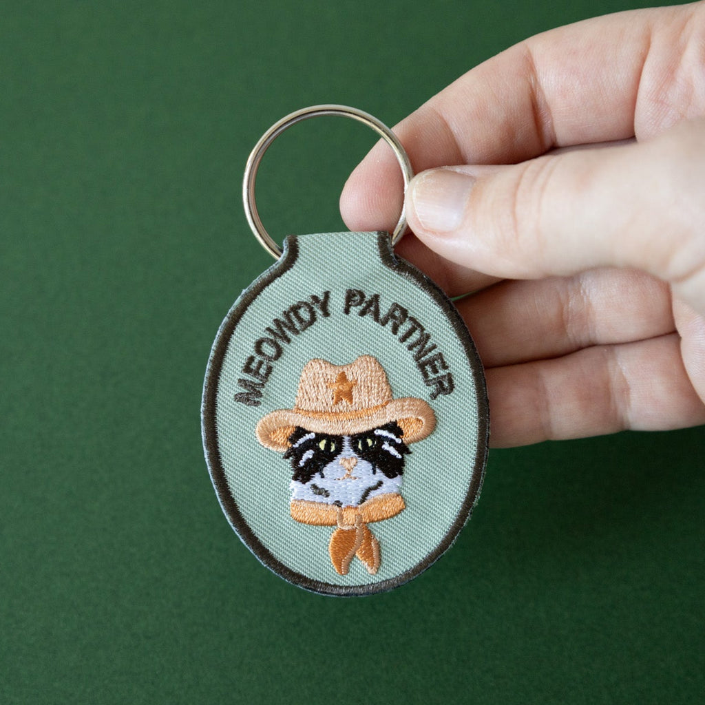 A hand holding a green embroidered keychain with a cat wearing a cowboy hat and the text 'meowdy parner' held over a green background.