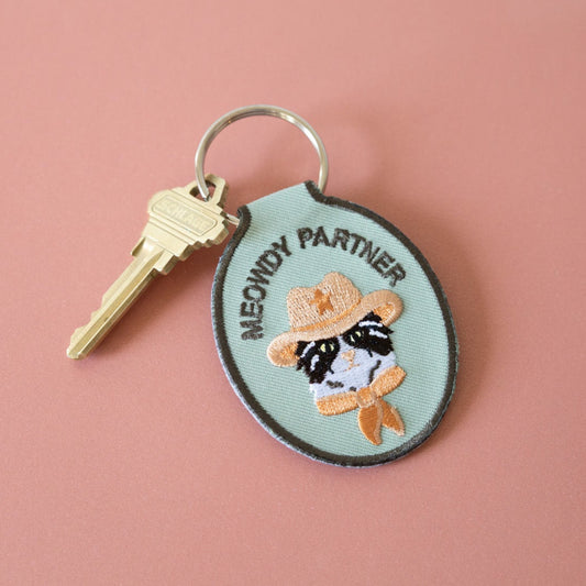 A green embroidered keychain with a design of a cat wearing a cowboy hat and the text 'MEOWDY PARTNER'. There is a metal key attached to it.