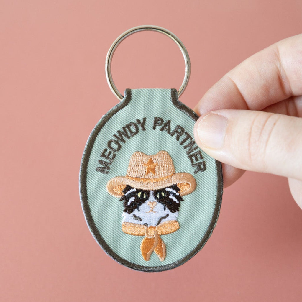 A hand holding a green embroidered keychain with a cat wearing a cowboy hat and the text 'meowdy parner' held over a pink background.