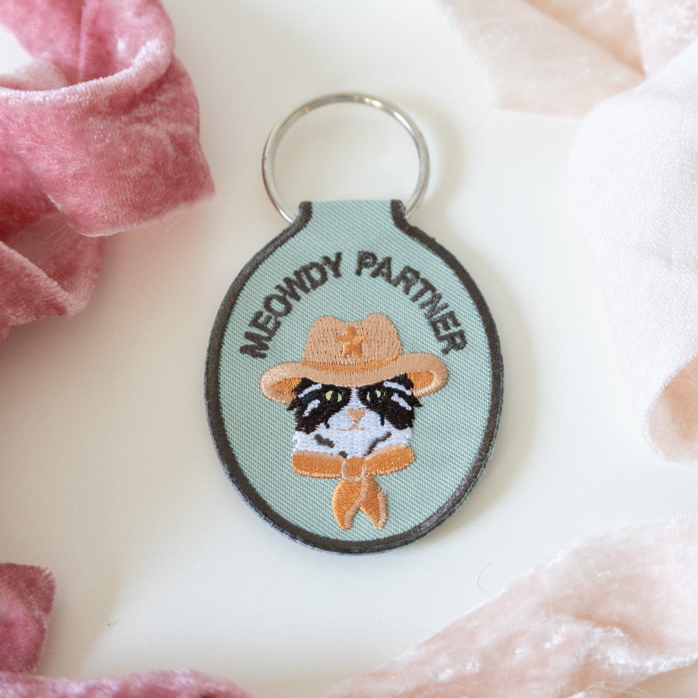 A green embroidered keychain with a cat wearing a cowboy hat and the text 'meowdy partner' displayed on a white background with pink ribbons.