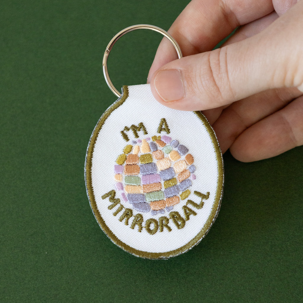 A hand holding white twill keychain with an embroidered disco ball design and the text 'I'm a mirrorball" displayed on a green background.