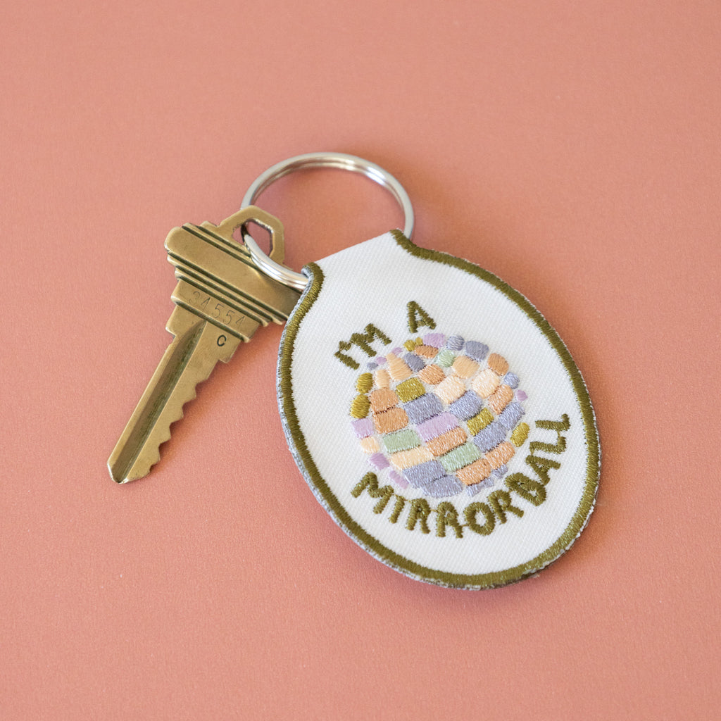 A white twill keychain with an embroidered disco ball design and the text 'I'm a mirrorball" displayed on a pink background.