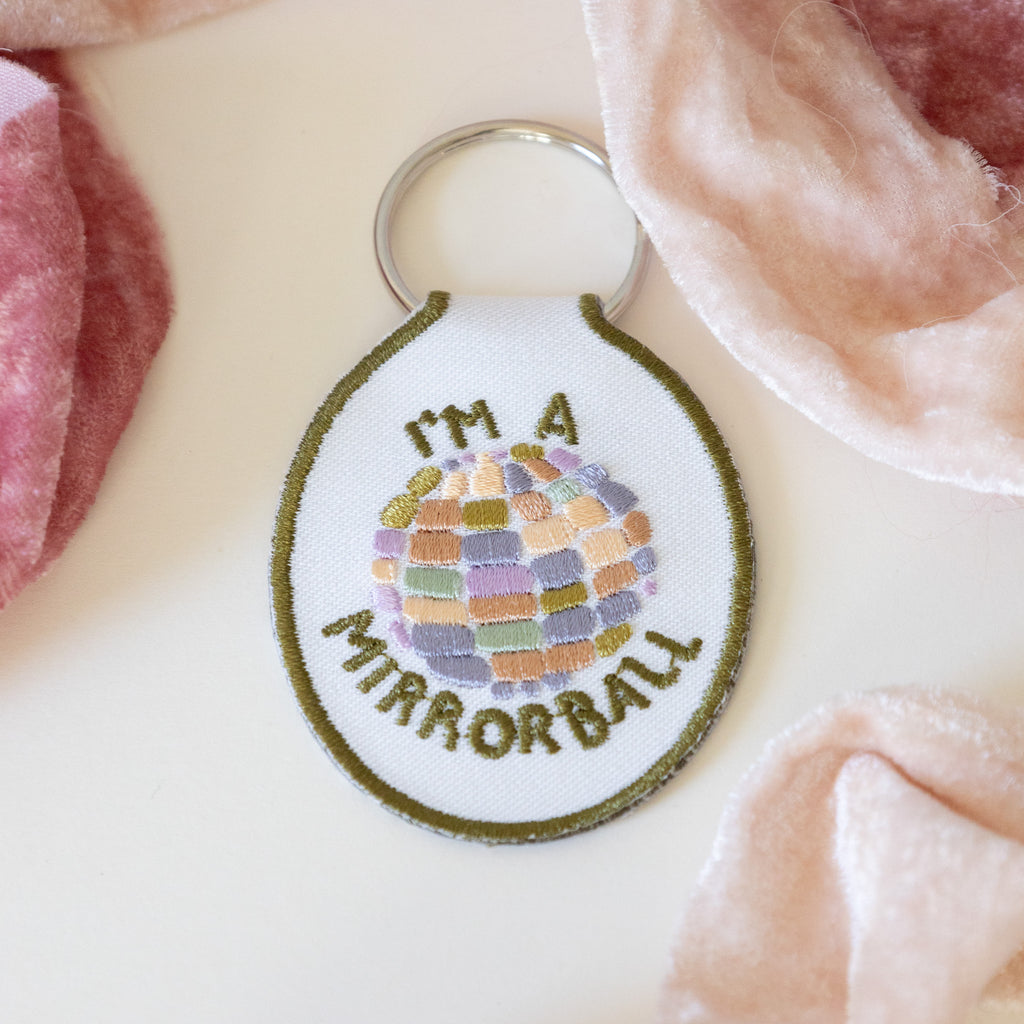 A white twill keychain with an embroidered disco ball design and the text 'I'm a mirrorball" displayed on a white background and pink ribbons.