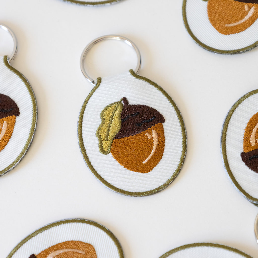 Multiple white twill keychains with an acorn embroidered design, displayed on a white background.