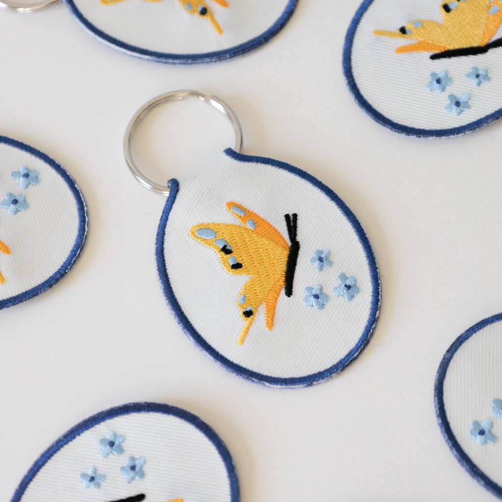 Multiple white twill keychains with an embroidered yellow butterfly and blue flowers, displayed on a white background.