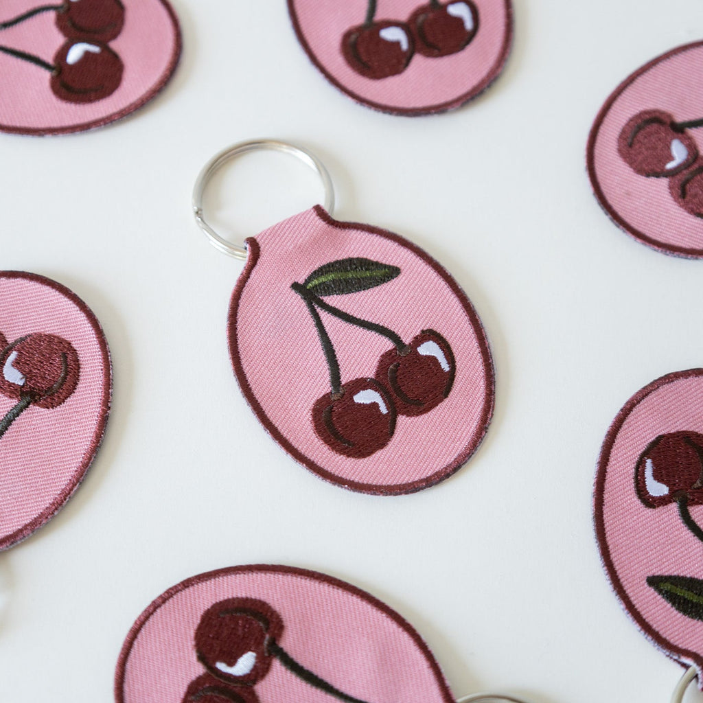 Embroidered keychains with a cherry embroidery design on a pink cotton twill patch, attached to a metal split key ring, displayed on a white background.