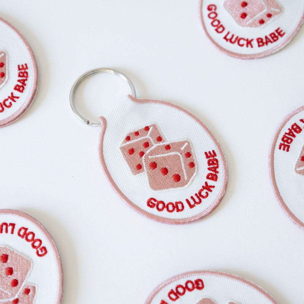 Multiple white cotton twill keychains with red and pink embroidered dice and the text 'GOOD LUCK BABE' displayed on a white background.