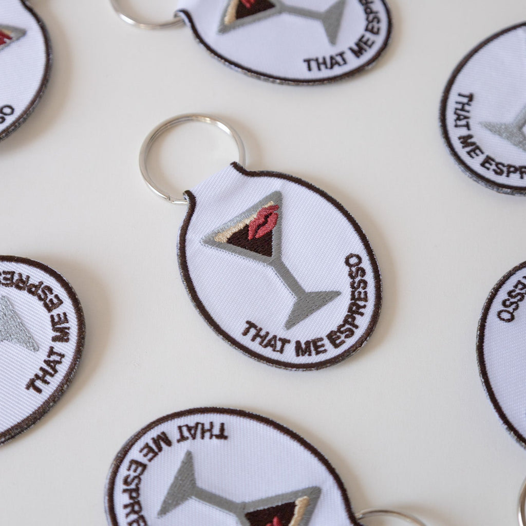 Embroidered keychains with a design of an espresso martini with a kiss and the phrase 'that me espresso' on it, attached to a metal key ring, displayed on a white background.