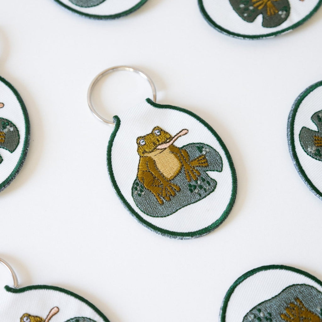 Multiple cream twill keychains with an embroidered frog on a lily pad design, displayed against a white background.