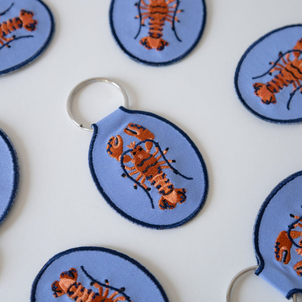 Multiple blue keychains with an embroidered red and blue lobster design, displayed against a white background.