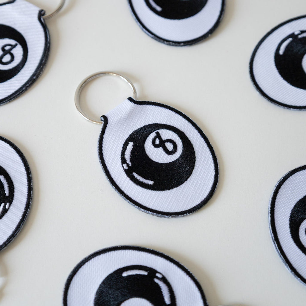 Multiple embroidered keychains with a white background featuring a black eight ball design, displayed on a white background.