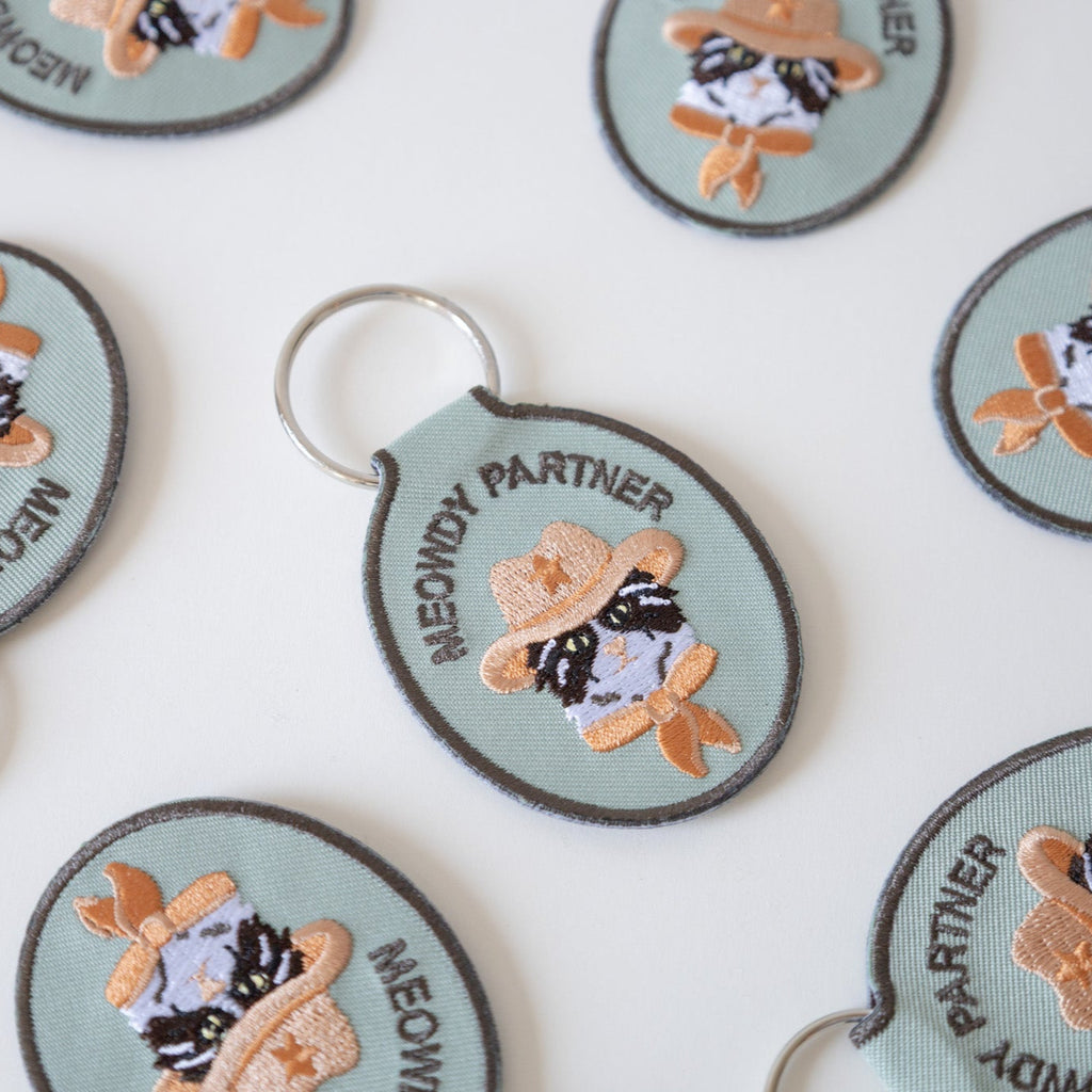 Green embroidered keychains with cats wearing cowboy hats and text that says 'meowdy partner' arranged on a white background.