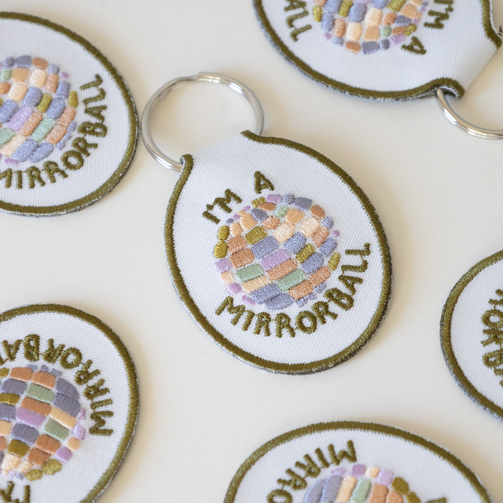 Multiple white twill keychains with an embroidered disco ball design and the text 'I'm a mirrorball" displayed on a pink background.