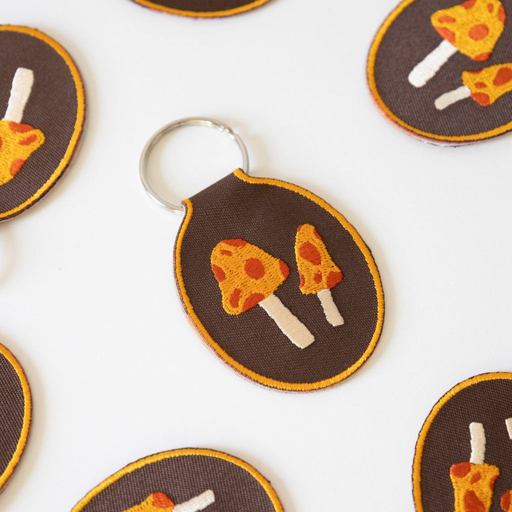 Multiple brown embroidered keychains featuring two yellow and red mushrooms displayed on a white background.