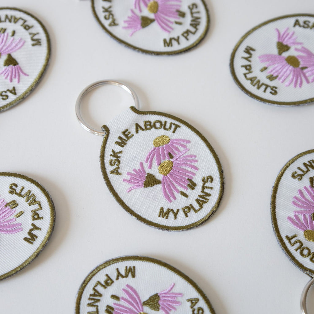 Multiple keychains with a cream cotton twill patch embroidered with the text 'ASK ME ABOUT MY PLANTS' and a floral design, displayed against a white background.