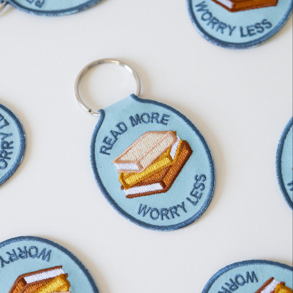Multiple blue embroidered keychains with stacked books and the text "read more worry less" displayed on a white background.