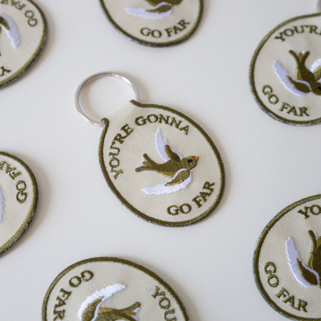 Multiple beige keychains with the embroidered text 'YOU'RE GONNA GO FAR' and an embroidered bird illustration on them, displayed against a white background.