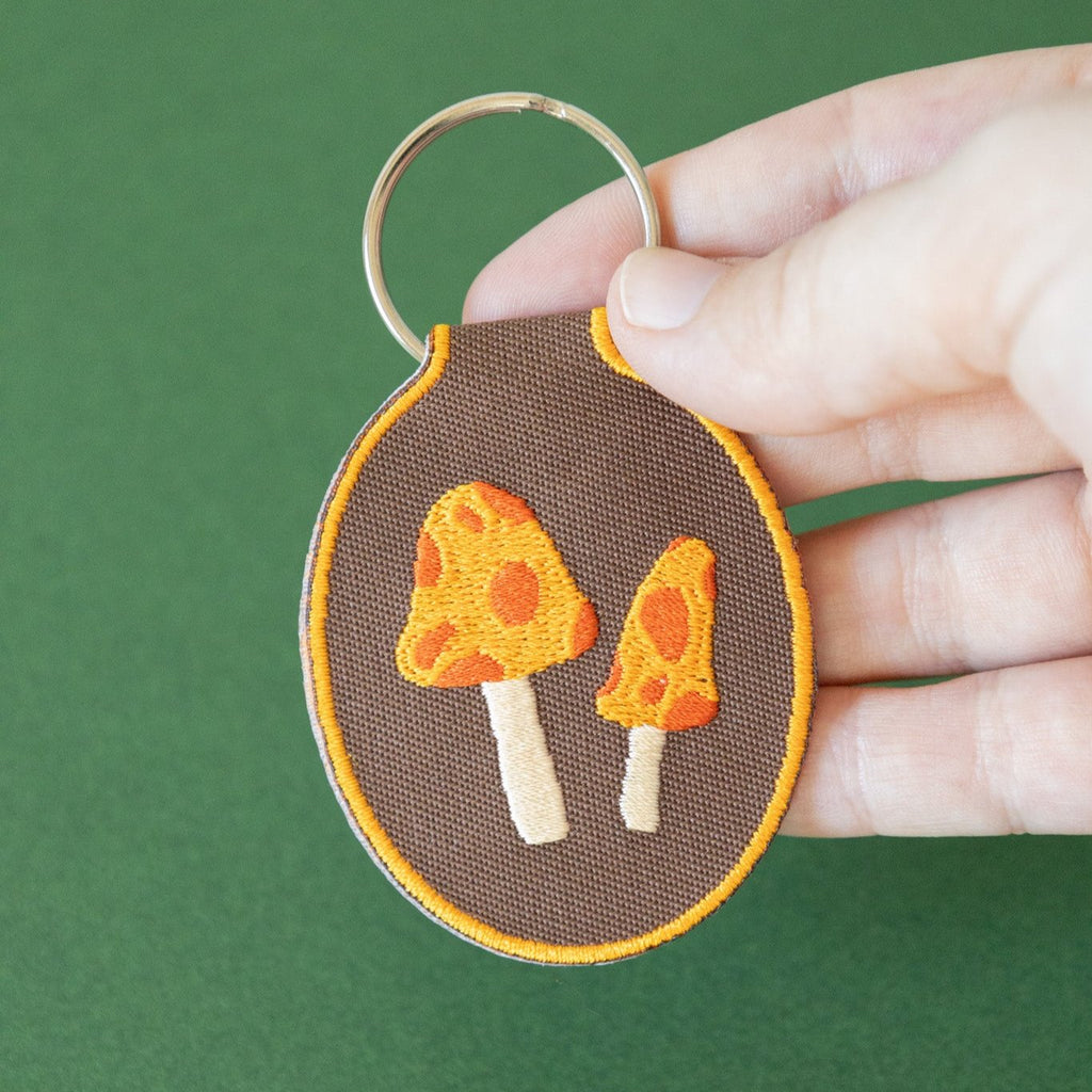 A hand holding a brown embroidered keychain featuring two yellow and red mushrooms displayed on a green background.