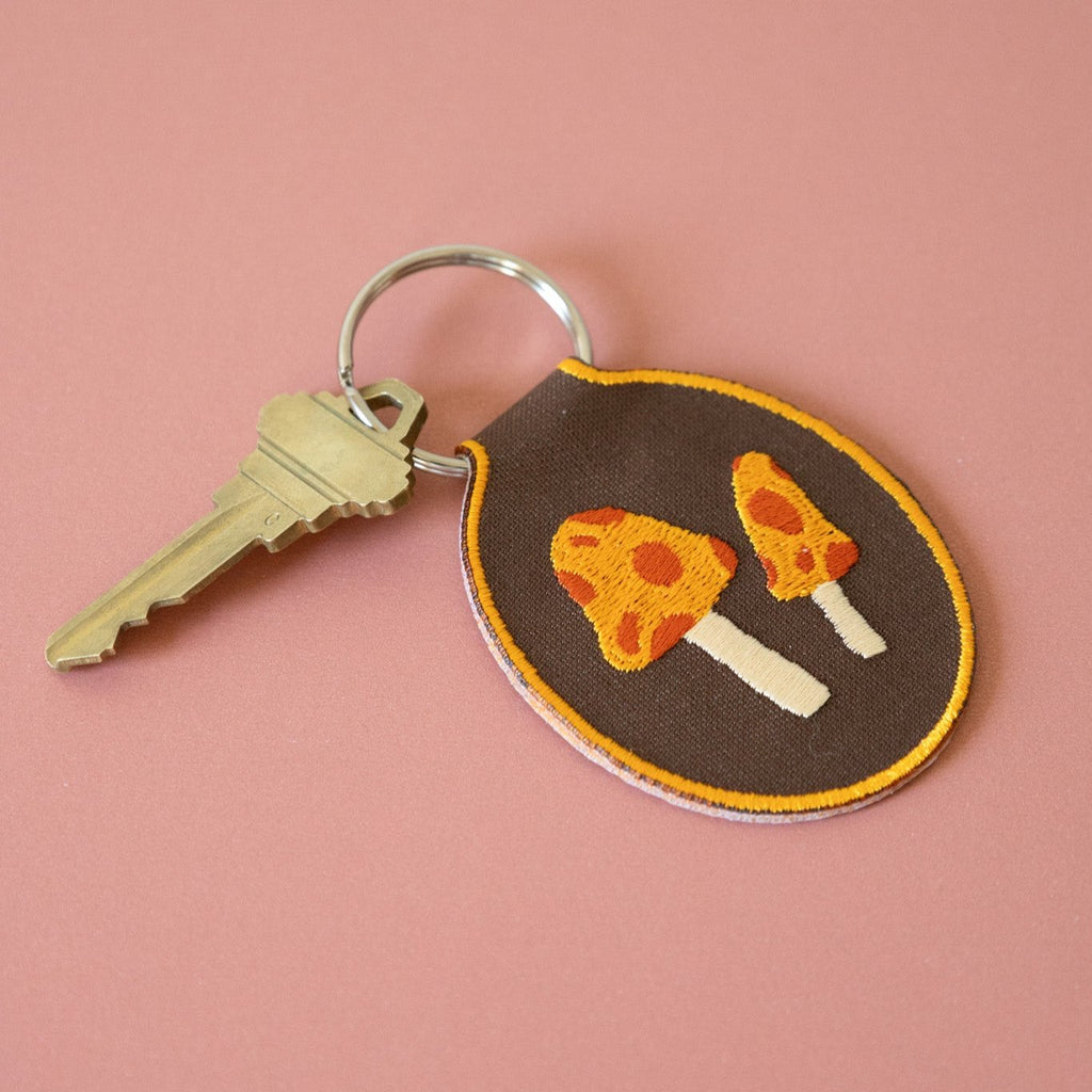 A brown embroidered keychain featuring two yellow and red mushrooms displayed on a pink background.