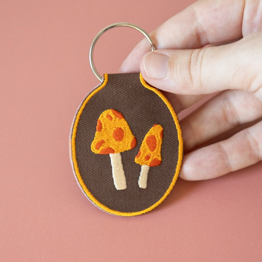 A hand holding a brown embroidered keychain featuring two yellow and red mushrooms displayed on a pink background.