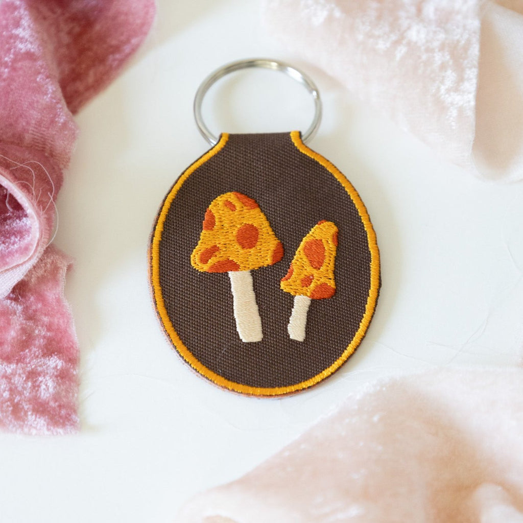 A brown embroidered keychain featuring two yellow and red mushrooms displayed on a white background with pink ribbons.