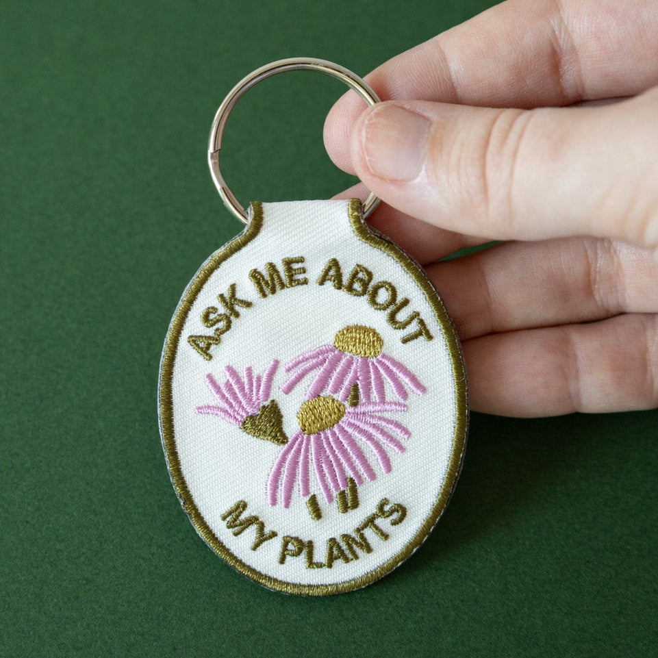 A hand holding a keychain with a cream cotton twill patch embroidered with the text 'ASK ME ABOUT MY PLANTS' and a floral design, displayed against a green background.