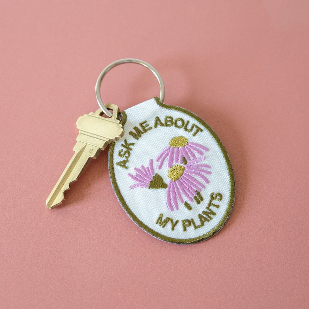 A keychain with a cream cotton twill patch embroidered with the text 'ASK ME ABOUT MY PLANTS' and a floral design, displayed against a pink background.