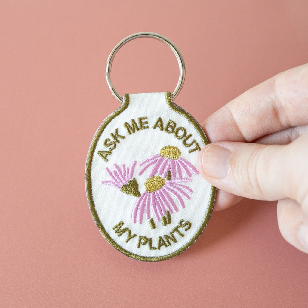 A hand holding a keychain with a cream cotton twill patch embroidered with the text 'ASK ME ABOUT MY PLANTS' and a floral design, displayed against a pink background.