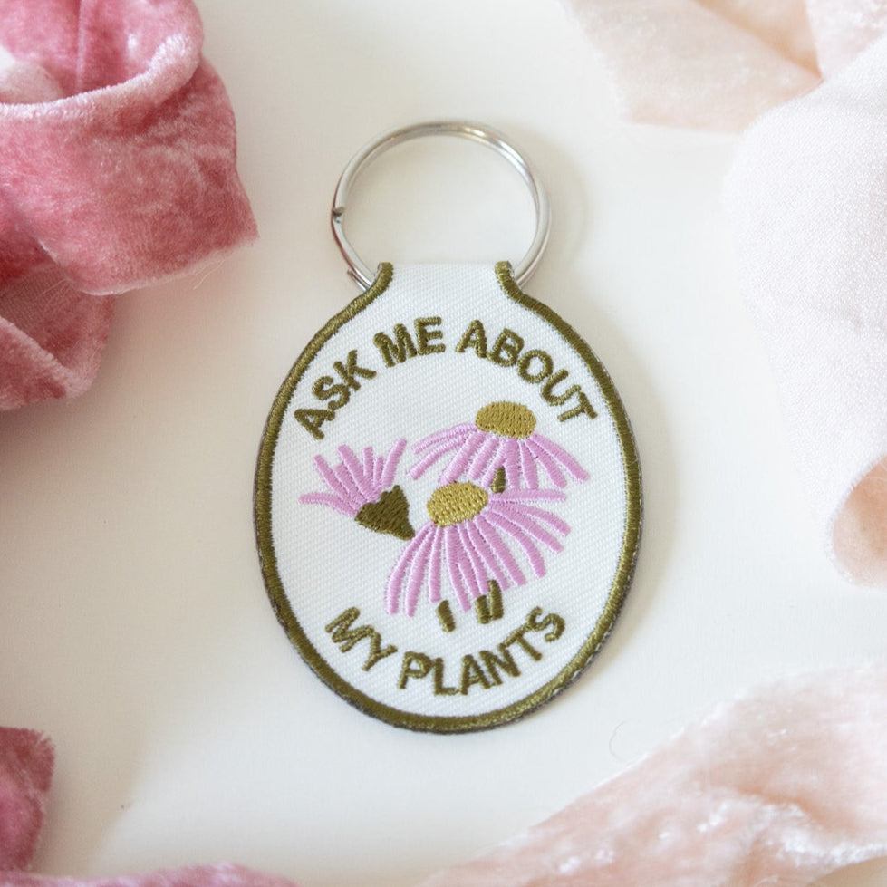 A keychain with a cream cotton twill patch embroidered with the text 'ASK ME ABOUT MY PLANTS' and a floral design, displayed against a white background with pink ribbons.