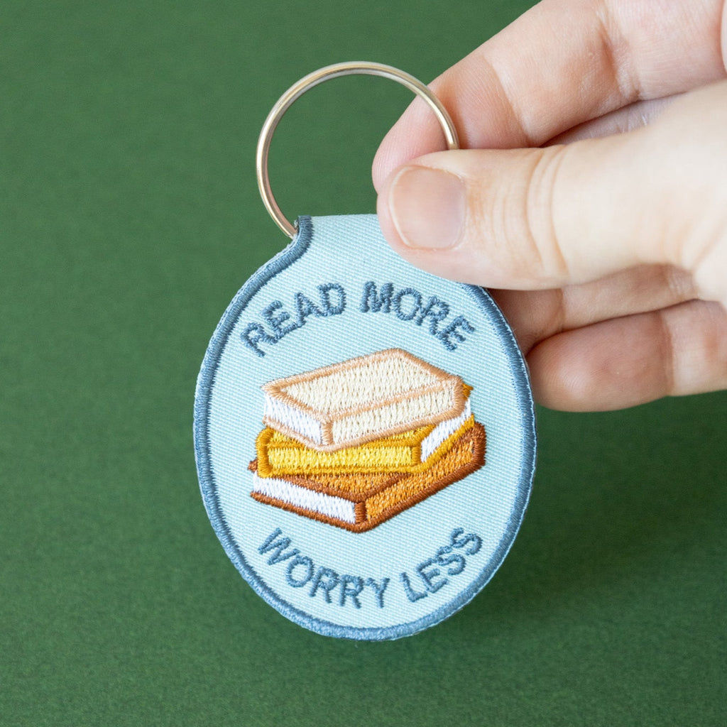 A hand holding a blue embroidered keychain with stacked books and the text "read more worry less" displayed on a green background.