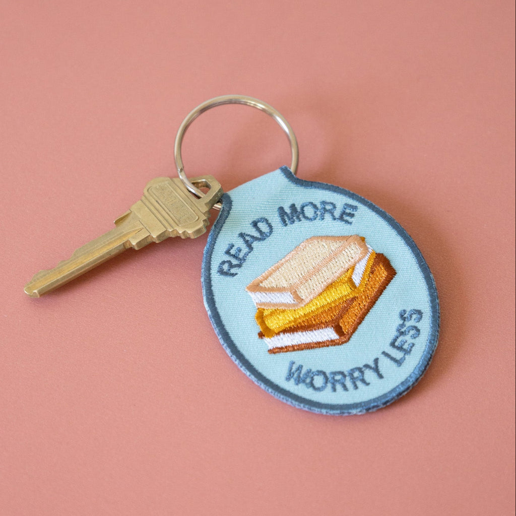 A blue embroidered keychain with stacked books and the text "read more worry less" displayed on a pink background.