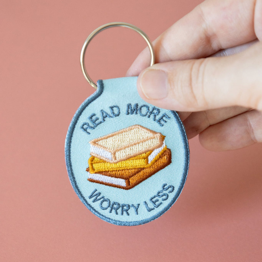A hand holding a blue embroidered keychain with stacked books and the text "read more worry less" displayed on a pink background.