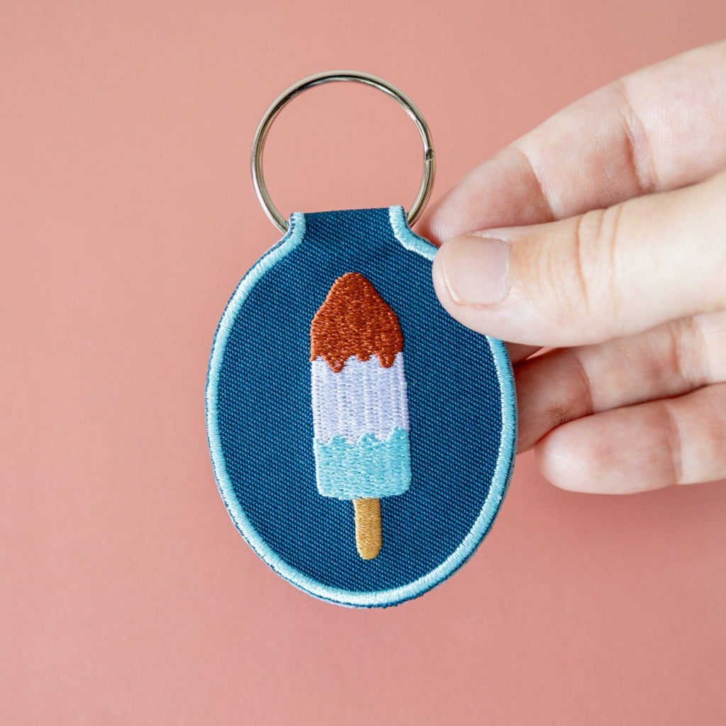 Embroidered keychain with red, white, and blue ice pop design held by a hand against a pink background