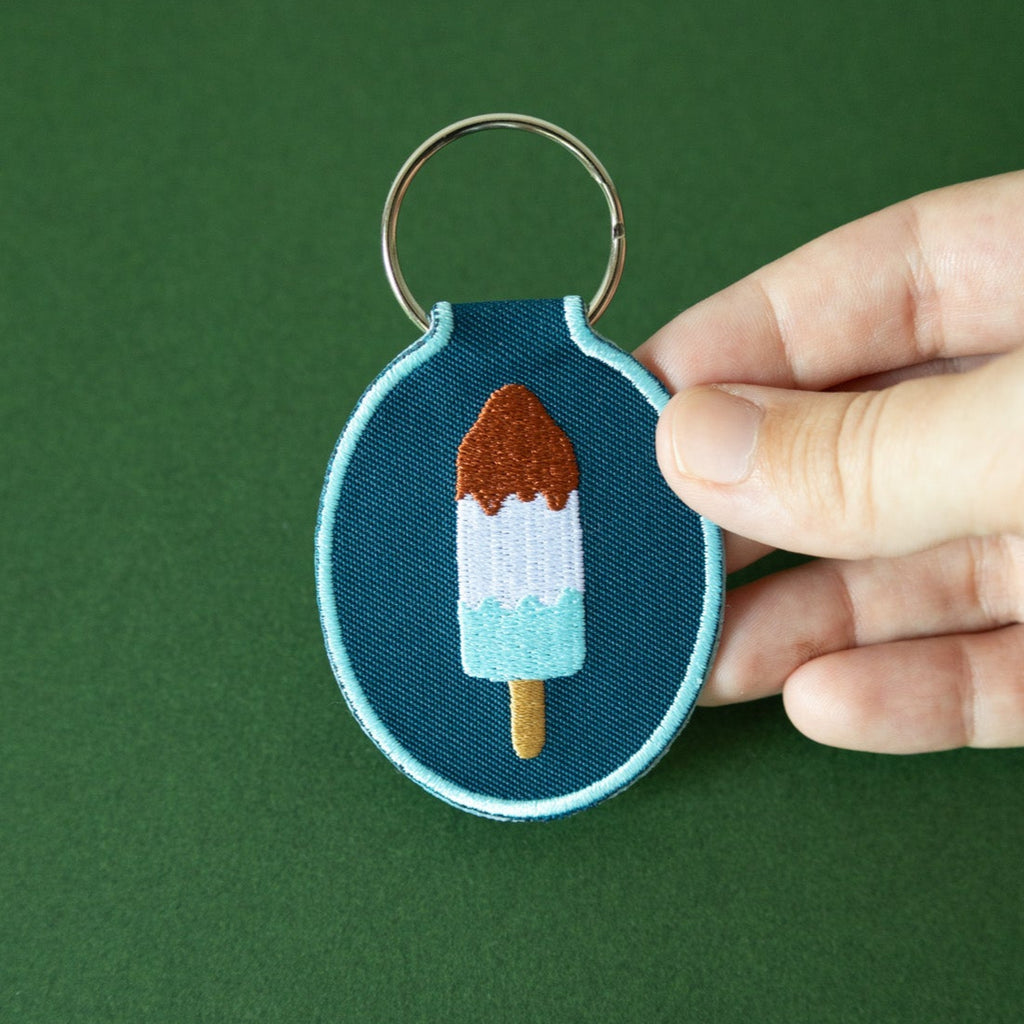 Hand holding an embroidered keychain with a red, white, and blue ice pop design against a green background