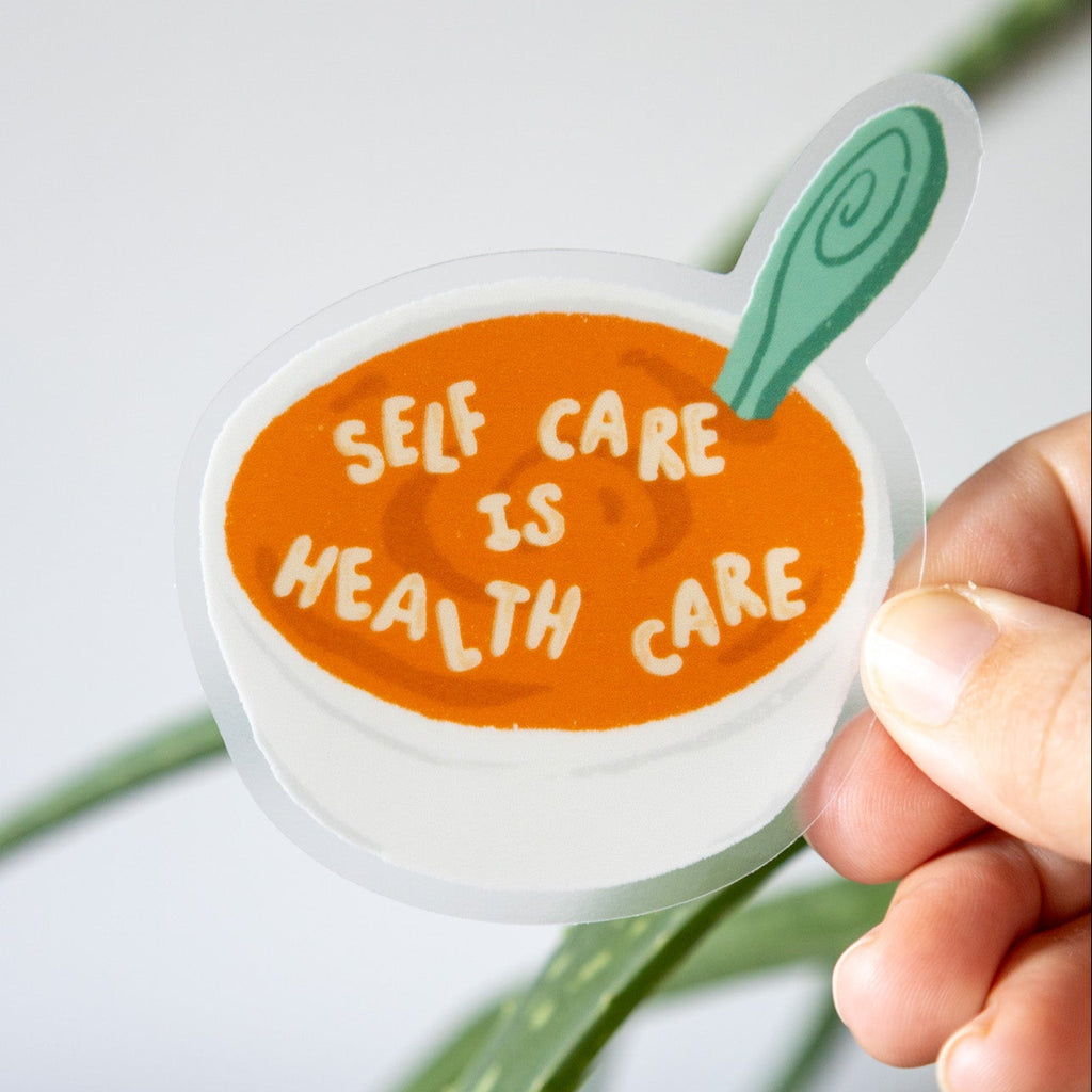 Sticker with 'Self care is health care' text held by a hand with a plant in the background