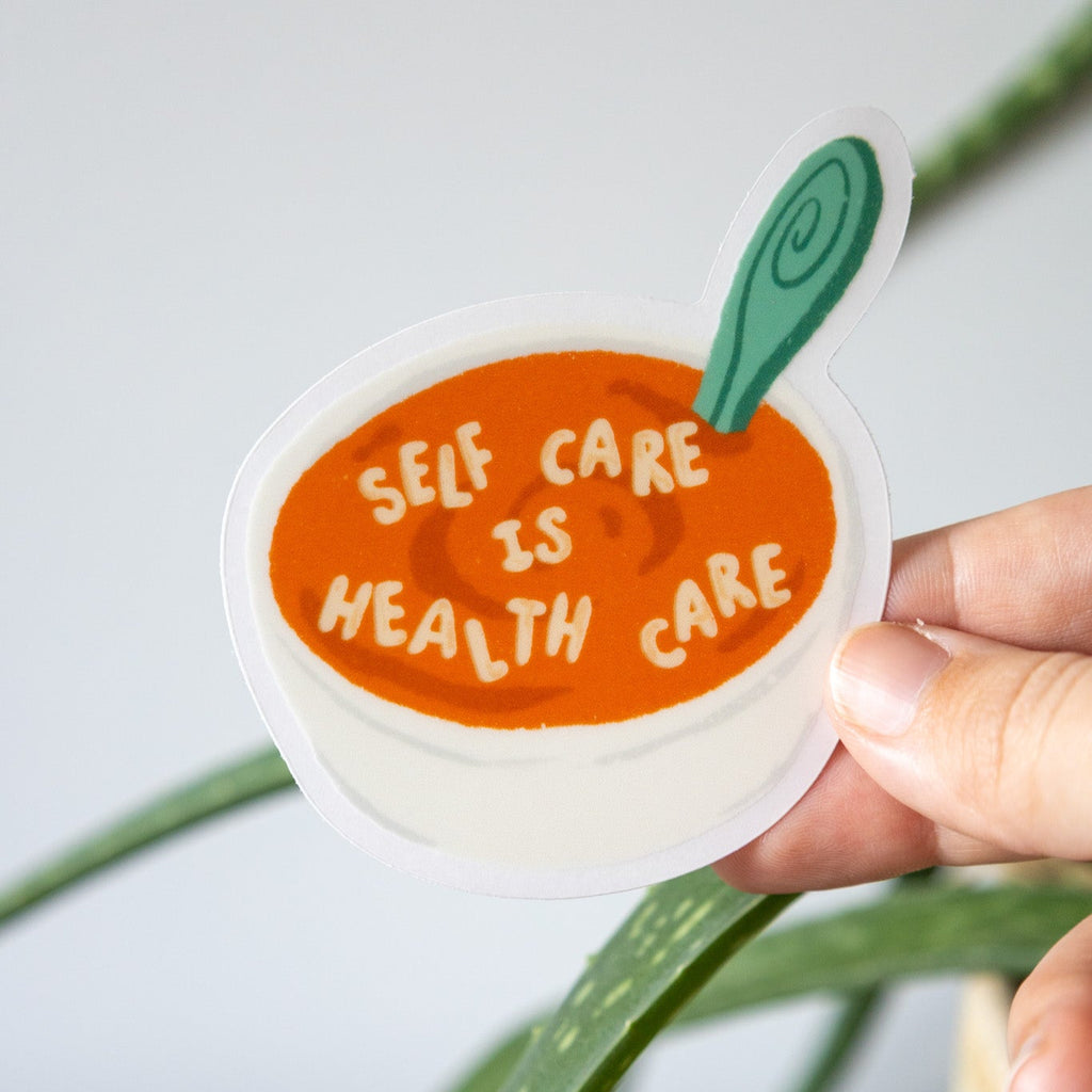 Sticker with 'Self care is health care' text held by a hand against a light background