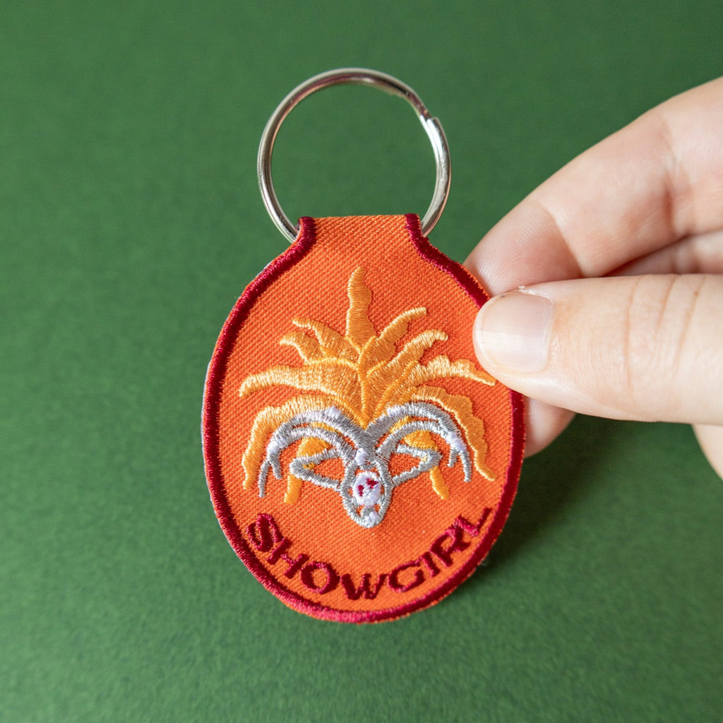 Hand holding an orange keychain with 'Showgirl' design on a green background