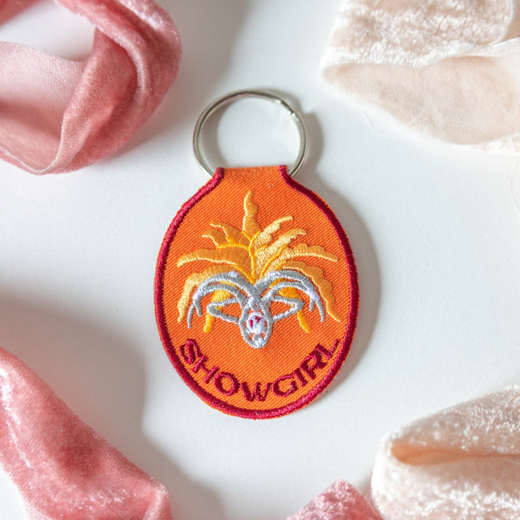 Orange embroidered keychain with 'Showgirl' design on a white background with pink ribbons.