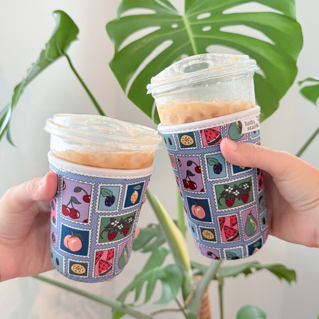 Two hands holding coffee cups with patterned sleeves in front of a plant.