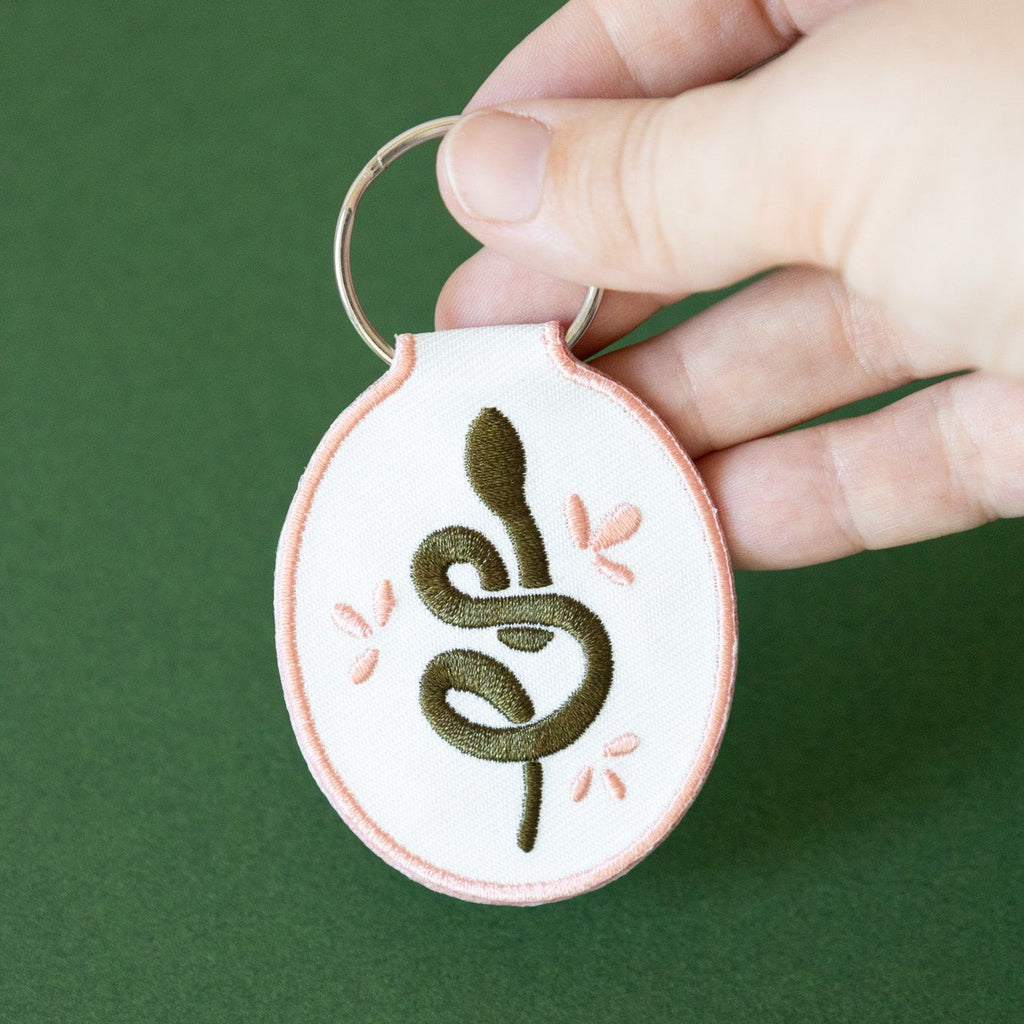 A hand holding a white cotton twill keychain with an embroidered snake design, displayed on a green background.